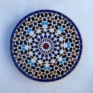 May include: A round decorative plate with a geometric pattern in shades of blue, brown, white, and red. The pattern is reminiscent of traditional Moroccan tilework.