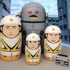 May include: A set of four nesting dolls, or matryoshka dolls, featuring a robot and three people wearing yellow and white safety vests and helmets. The robot is gray and has red eyes. The dolls are painted in a realistic style. A silver boombox is also included in the image.