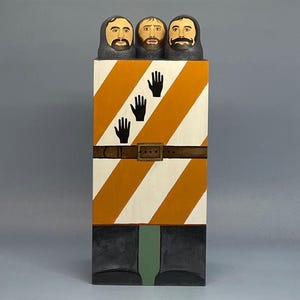 Puede incluir: Escultura de arte popular de madera con tres caras barbadas en la parte superior. El cuerpo está pintado con rayas diagonales naranjas y blancas, manos negras, un cinturón marrón y botas negras. Fondo gris. Escultura única.