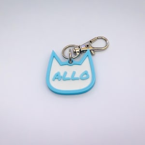 May include: A cat-shaped keychain with a light blue border and the word "ALLO" in light blue. The keychain has a silver clasp and a small ring for attaching keys. The background is white.