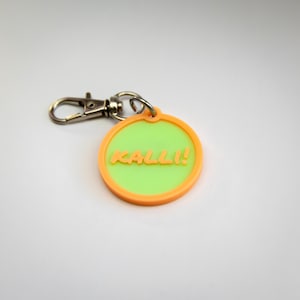 May include: A round keychain with a lime green center and an orange border. The text "KALLI!" is in the center. The keychain has a silver-colored clasp and ring.