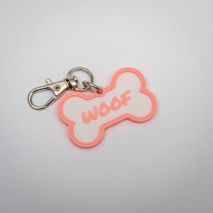 May include: A dog bone-shaped pet tag with the word "WOOF" in raised letters. The tag is white with a pink border and includes a silver-colored clasp for easy attachment to a collar.