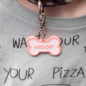 May include: A close-up of a dog tag in the shape of a bone, with a pink border and the word "WOOF" in the center. The tag is attached to a silver clasp and a brown leather collar. The background is a light blue t-shirt with black text.