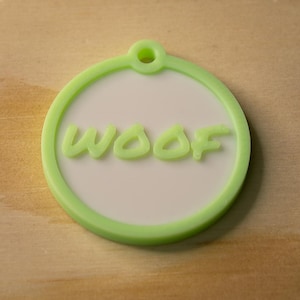 May include: A round, light green pet tag with the word "WOOF" in a matching green, raised font. The tag has a white center and a hole at the top for attachment. The tag is on a light brown wooden surface.