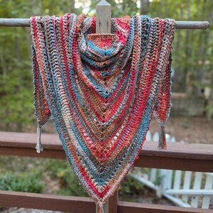 May include: A triangular crochet shawl featuring a variegated yarn pattern in shades of red, blue, brown, and cream. The garment has a textured stitch design with a pointed bottom edge and three small tassels attached to the corners and center point.
