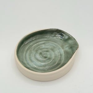 May include: A ceramic spoon rest with a light green interior and a cream-colored exterior. The interior has a spiral design. The spoon rest has a small spout on one side.