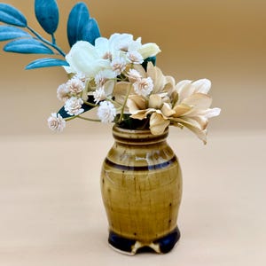 May include: A small, glossy, olive-green ceramic vase with a dark brown base and rim, containing an arrangement of artificial flowers. The bouquet features teal-colored leaves, white blossoms, and light tan petals, creating a contrast in color.