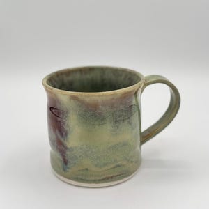 May include: Handmade ceramic mug with a green, burgundy, and cream marbled glaze. The mug has a rounded body, a sturdy handle, and a white base. This unique mug is perfect for coffee, tea, or any hot beverage.