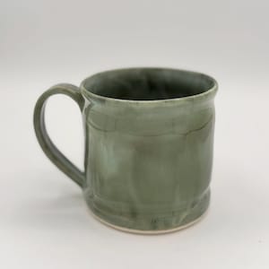 May include: A handmade ceramic mug with a rounded body and a curved handle. The mug is glazed in a mottled green color, with variations in tone. The base of the mug is a lighter, off-white color. The mug is designed for hot or cold beverages.