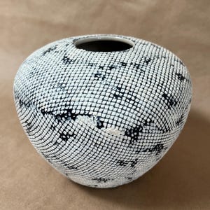 May include: A round ceramic vase with a textured surface. The vase is primarily white with a pattern of small black dots, creating a grid-like design. The opening is black.