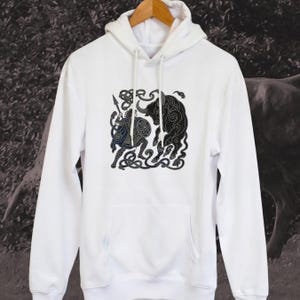 May include: A white hooded sweatshirt featuring a black graphic print on the chest. The design depicts a figure holding a spear and shield facing a bull, both rendered in a stylized, swirling line art pattern with intricate knotwork details.