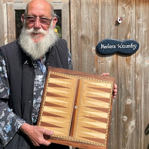 Puede incluir: Un tablero de backgammon de madera con incrustaciones de madera claras y oscuras. La persona sostiene el tablero frente a un cobertizo de madera. El letrero en el cobertizo dice "Merlin's Wizardry".