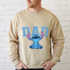 May include: A beige crewneck sweatshirt featuring the word DAD in large light blue block letters. Centered below the text is a graphic illustration of the blue character Stitch with large ears, sitting in a forward-facing position on the chest.