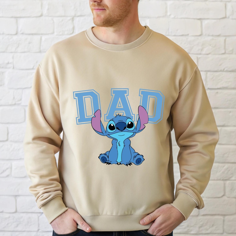 May include: A beige crewneck sweatshirt featuring the word DAD in large light blue block letters. Centered below the text is a graphic illustration of the blue character Stitch with large ears, sitting in a forward-facing position on the chest.