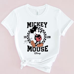 May include: White t-shirt featuring a black and white graphic of Mickey Mouse in a chequered circle. The text "Mickey Mouse" is written in black above the graphic and "Disney" is written in black below the graphic. The numbers "19" and "28" are written on either side of the circle.