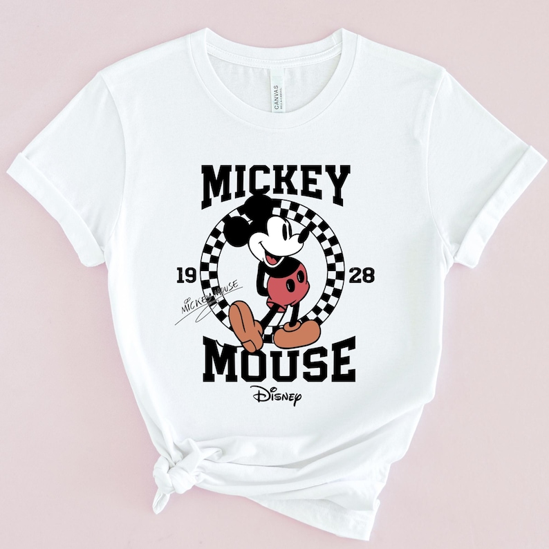 May include: White t-shirt featuring a black and white graphic of Mickey Mouse in a chequered circle. The text "Mickey Mouse" is written in black above the graphic and "Disney" is written in black below the graphic. The numbers "19" and "28" are written on either side of the circle.