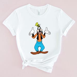 May include: White t-shirt with a cartoon image of Goofy, a Disney character, with his arms outstretched. Goofy is wearing a yellow hat, orange shirt, blue pants, and brown shoes.