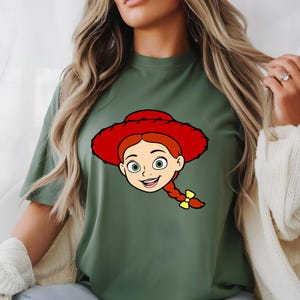 May include: Olive green t-shirt featuring a cartoon illustration of a character with red hair, a red hat, and a yellow bow. The character has green eyes and a smiling expression. The t-shirt is a casual style.