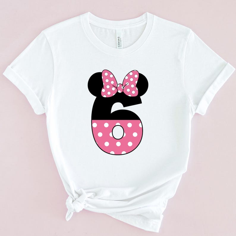 May include: White t-shirt featuring a black number six with a pink polka-dotted design. The number six is topped with black mouse ears and a pink polka-dotted bow. The shirt is made of a soft material.