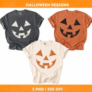 Peut inclure: Trois t-shirts à manches courtes et col rond avec un motif de visage de citrouille d'Halloween vieilli. Un t-shirt est gris anthracite avec une impression blanche, un orange avec une impression noire, et un crème avec une impression orange. Texte HALLOWEEN DESIGNS.