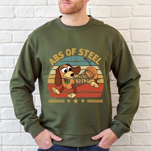 Puede incluir: Sudadera verde oliva con un gr&aacute;fico de un perro salchicha de juguete de dibujos animados con un cuerpo de resorte, el texto "ABS OF STEEL" y un dise&ntilde;o retro de puesta de sol. El dise&ntilde;o incluye estrellas y rayas.