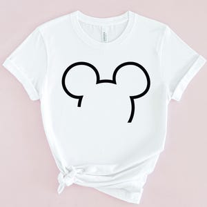 Op de afbeelding: Wit T-shirt met een zwarte omtrek van een muizenkop. Het T-shirt heeft korte mouwen en een ronde hals. De onderkant van het T-shirt is geknoopt. De achtergrond is lichtroze.