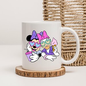 May include: White ceramic mug with a colorful cartoon image of Minnie Mouse and Daisy Duck wearing heart-shaped sunglasses. Minnie is holding a pink and blue drink and Daisy is holding an ice cream cone.