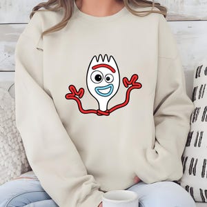 May include: A beige sweatshirt featuring a cartoon image of Forky from Toy Story. The graphic shows Forky's white fork body, red arms, and a smiling face with a blue mouth and one eye. The sweatshirt is a casual, long-sleeved garment.