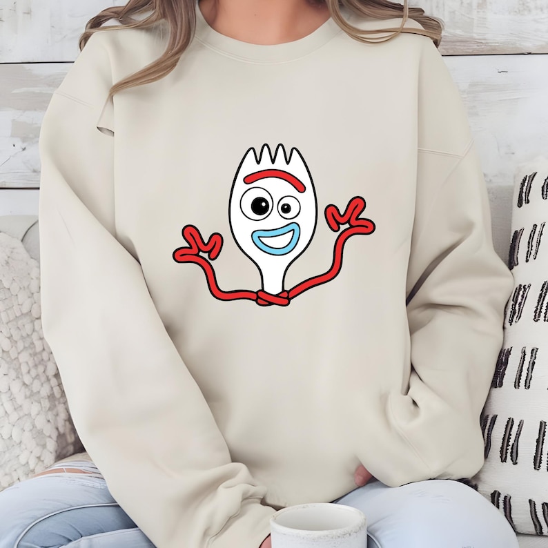 May include: A beige sweatshirt featuring a cartoon image of Forky from Toy Story. The graphic shows Forky's white fork body, red arms, and a smiling face with a blue mouth and one eye. The sweatshirt is a casual, long-sleeved garment.