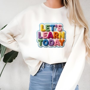 May include: Cream-colored sweatshirt with a colorful, embroidered patch. The patch features the text "LET'S LEARN TODAY" in a variety of colors, including blue, pink, yellow, green, and orange. The sweatshirt is paired with blue jeans.