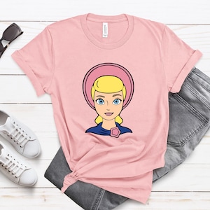May include: A light pink t-shirt featuring a cartoon illustration of a blonde woman with blue eyes wearing a pink bonnet and a blue dress. The shirt is paired with gray jeans and white sneakers.