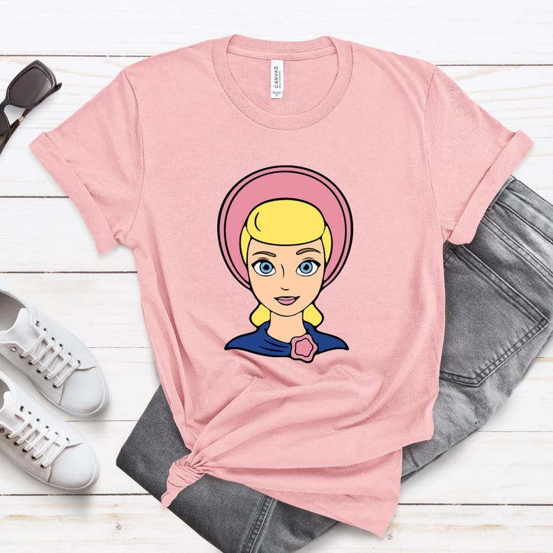 May include: A light pink t-shirt featuring a cartoon illustration of a blonde woman with blue eyes wearing a pink bonnet and a blue dress. The shirt is paired with gray jeans and white sneakers.