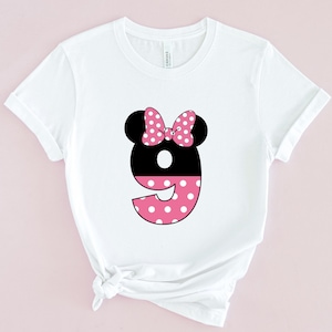 May include: White t-shirt featuring a black number nine with a pink polka dot design. The number has black mouse ears and a pink polka dot bow. The shirt is made of a soft material.