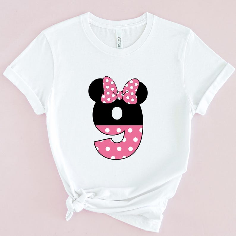 May include: White t-shirt featuring a black number nine with a pink polka dot design. The number has black mouse ears and a pink polka dot bow. The shirt is made of a soft material.