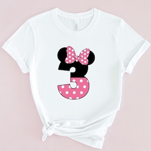 May include: White t-shirt featuring a black Minnie Mouse head with a pink polka dot bow. The number 3 is designed with a black top and a pink polka dot bottom. The shirt is made of a soft material.