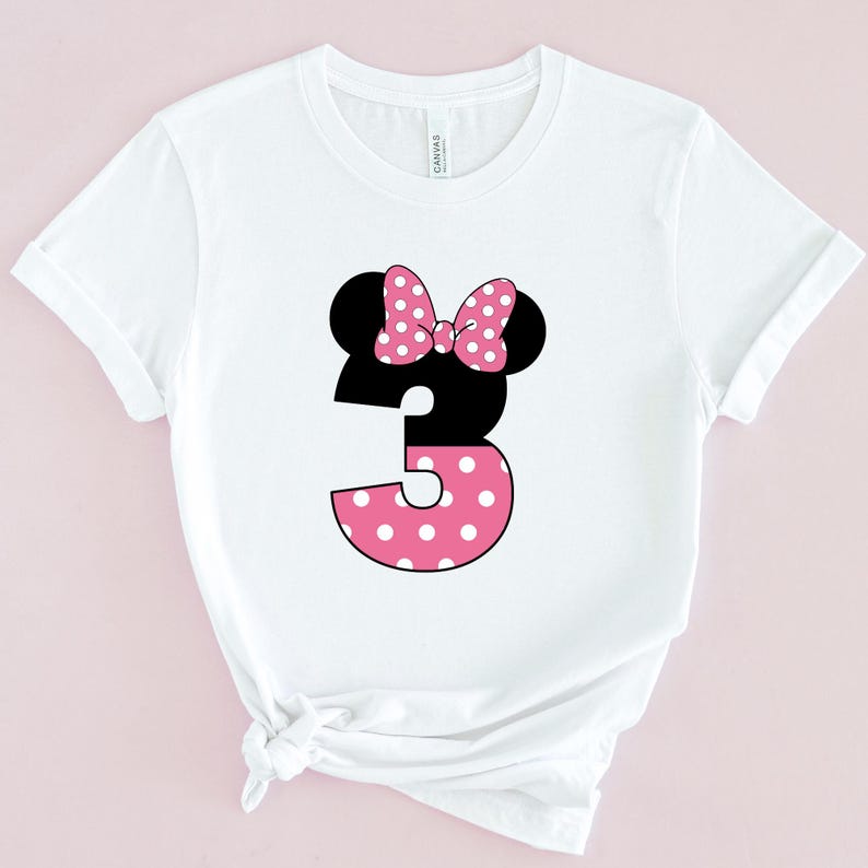May include: White t-shirt featuring a black Minnie Mouse head with a pink polka dot bow. The number 3 is designed with a black top and a pink polka dot bottom. The shirt is made of a soft material.