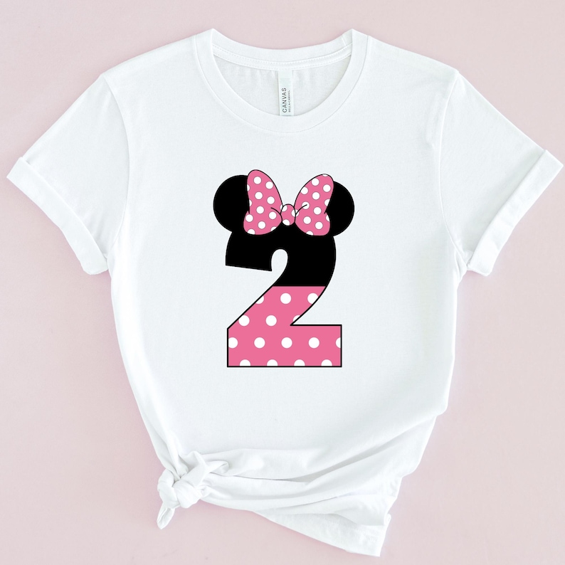 May include: White t-shirt featuring a black number 2 with a pink polka dot design. The number is topped with black mouse ears and a pink polka dot bow. The shirt is made of a soft material.