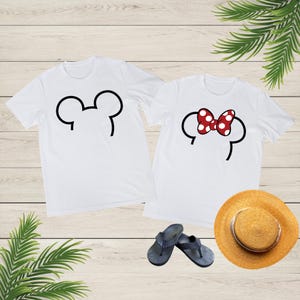 Op de afbeelding: Twee witte t-shirts met zwarte Mickey Mouse oorcontouren. E&eacute;n shirt heeft een simpele oorcontour, de andere een rood-witte polkadot strik. Een strohoed en donkerblauwe slippers staan ook in de afbeelding.