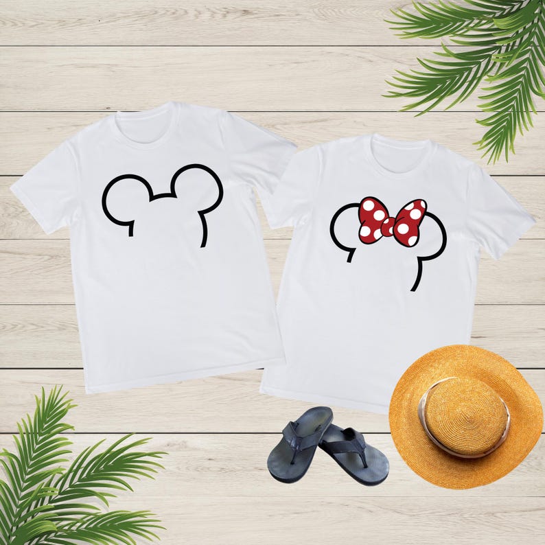 Op de afbeelding: Twee witte t-shirts met zwarte Mickey Mouse oorcontouren. E&eacute;n shirt heeft een simpele oorcontour, de andere een rood-witte polkadot strik. Een strohoed en donkerblauwe slippers staan ook in de afbeelding.