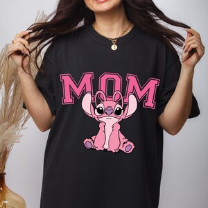 May include: A black t-shirt featuring the word MOM in large pink block letters with a pink graphic of the character Stitch from Lilo and Stitch positioned below the text. The character is depicted in a seated pose with large ears and dark eyes.