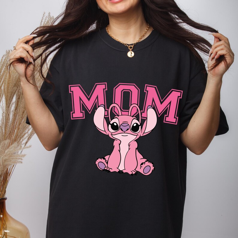 May include: A black t-shirt featuring the word MOM in large pink block letters with a pink graphic of the character Stitch from Lilo and Stitch positioned below the text. The character is depicted in a seated pose with large ears and dark eyes.