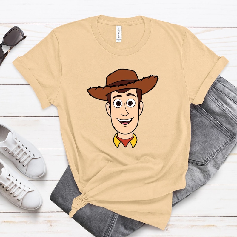 May include: A light tan t-shirt featuring a cartoon illustration of a cowboy character with a brown hat, brown hair, and a yellow shirt collar. The shirt is paired with gray jeans and white sneakers.