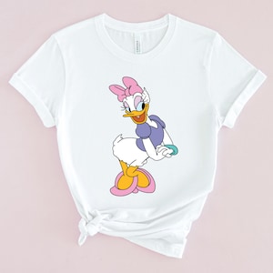 May include: White t-shirt with a cartoon image of a duck wearing a purple dress and a pink bow. The duck is standing on one leg and has a yellow beak and orange feet.