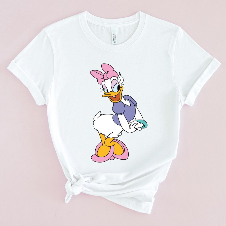 May include: White t-shirt with a cartoon image of a duck wearing a purple dress and a pink bow. The duck is standing on one leg and has a yellow beak and orange feet.