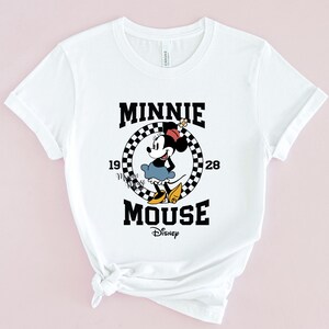 May include: White t-shirt with a black and white checkered circle graphic featuring Minnie Mouse in a red dress and yellow bow. The text "Minnie Mouse" is written in black with "1928" and "Disney" in smaller font.