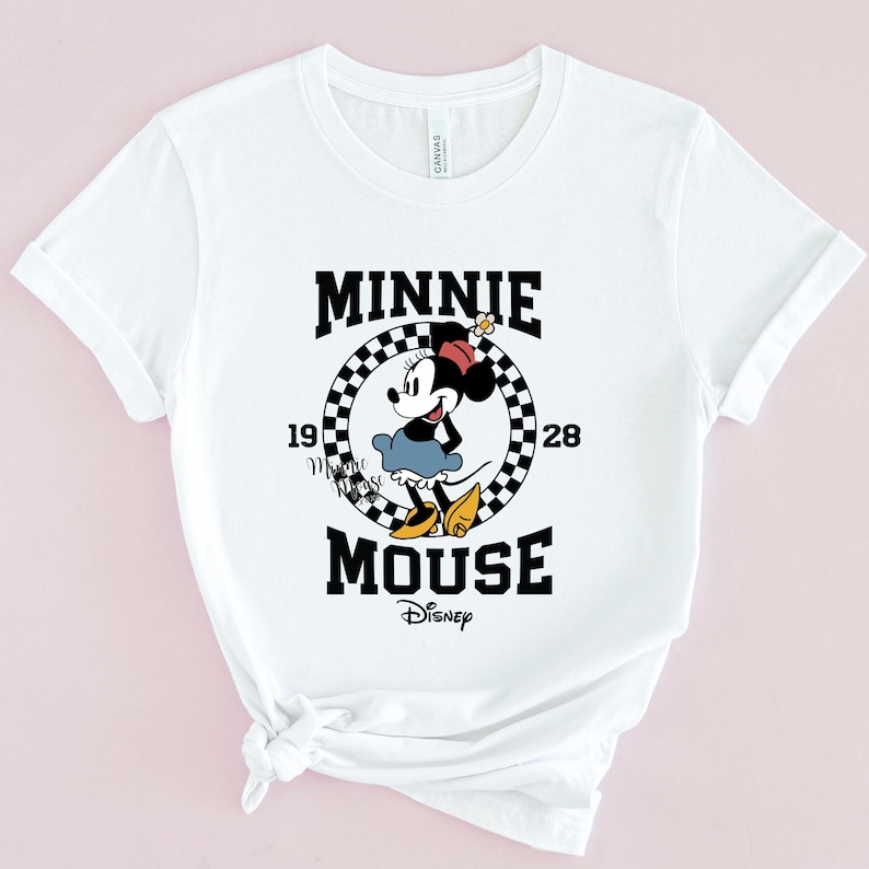 May include: White t-shirt with a black and white checkered circle graphic featuring Minnie Mouse in a red dress and yellow bow. The text "Minnie Mouse" is written in black with "1928" and "Disney" in smaller font.