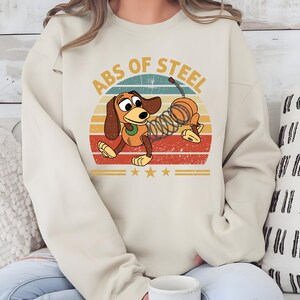 Puede incluir: Sudadera beige con un perro de dibujos animados con cuerpo de resorte, sobre un dise&ntilde;o retro de puesta de sol. El texto "ABS OF STEEL" est&aacute; arqueado sobre el perro. Tres estrellas doradas debajo.