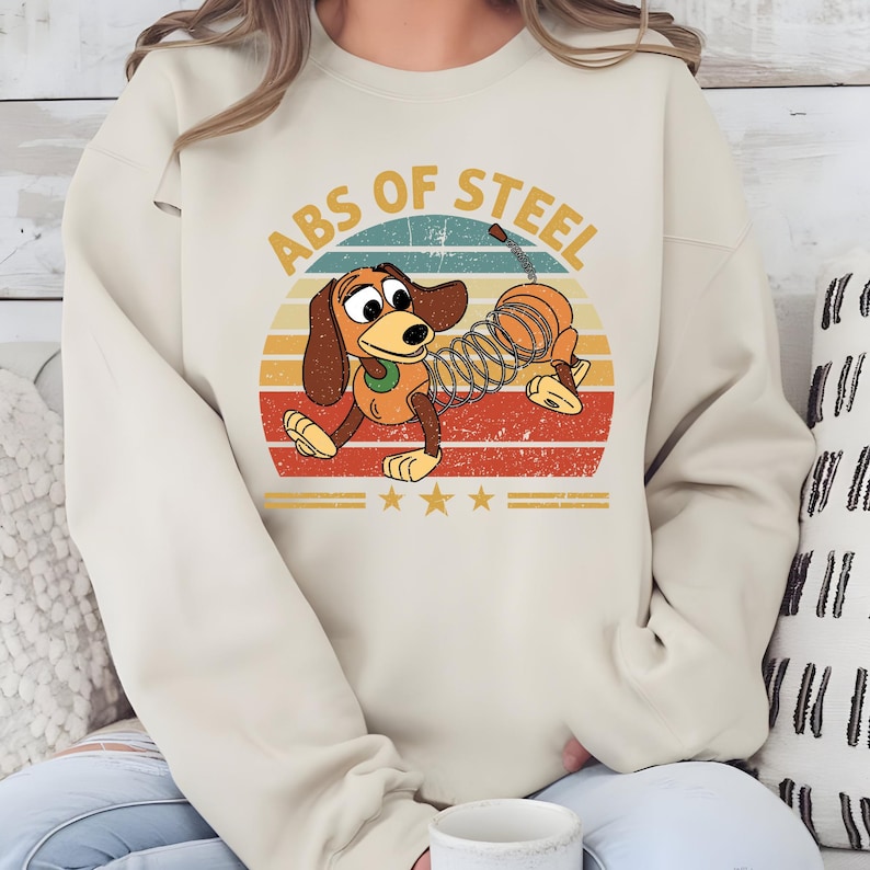 Puede incluir: Sudadera beige con un perro de dibujos animados con cuerpo de resorte, sobre un dise&ntilde;o retro de puesta de sol. El texto "ABS OF STEEL" est&aacute; arqueado sobre el perro. Tres estrellas doradas debajo.