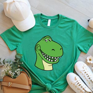 May include: Green t-shirt featuring a cartoon dinosaur head with a smiling expression. The dinosaur is green with a light yellow belly and white teeth. A white baseball cap and white sneakers are also visible.
