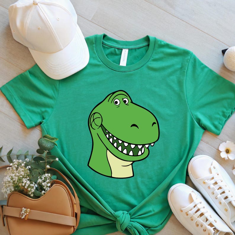 May include: Green t-shirt featuring a cartoon dinosaur head with a smiling expression. The dinosaur is green with a light yellow belly and white teeth. A white baseball cap and white sneakers are also visible.